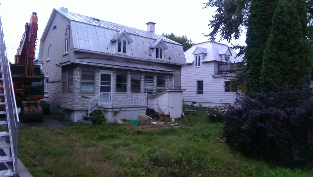 Une maison ancienne avec un toit en métal et une véranda, entourée de végétation. Une excavatrice est stationnée à l’arrière, suggérant une prochaine démolition.