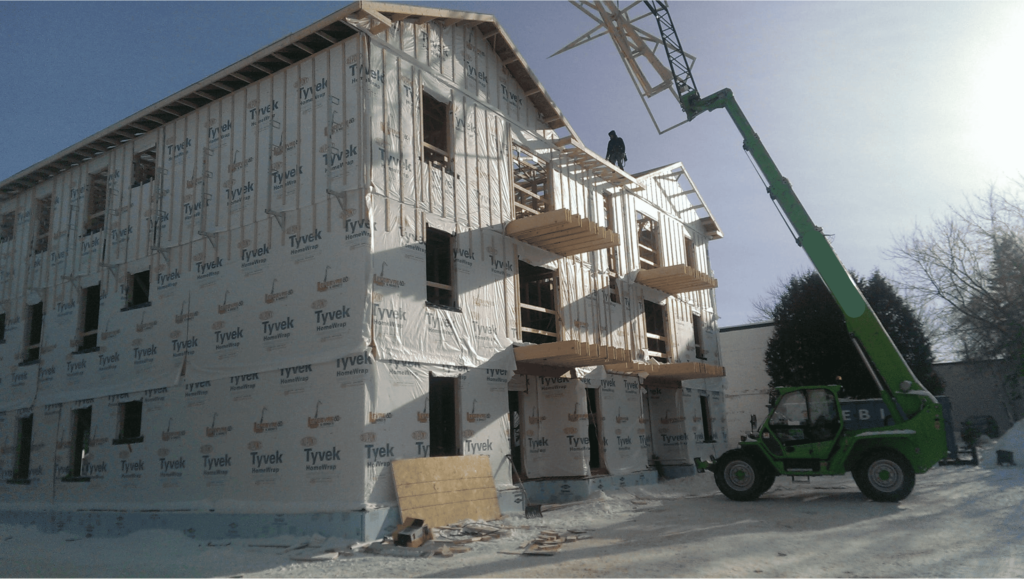 Travaux de construction d’un immeuble en ossature bois recouvert de Tyvek, avec un chariot télescopique vert soulevant une structure sous un ciel dégagé.