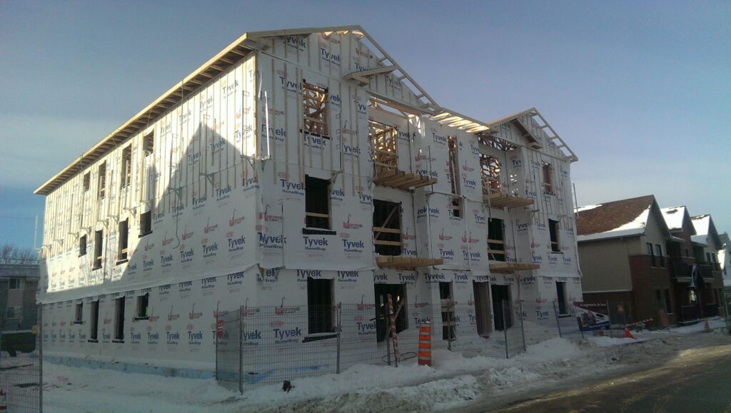 Bâtiment en construction avec structure en bois recouverte de pare-air Tyvek, entouré d’une clôture de chantier et de neige.