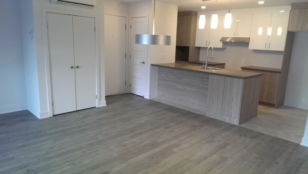 Intérieur d’un appartement moderne avec plancher en bois, cuisine ouverte avec îlot central et armoires en finition bois et blanc laqué.