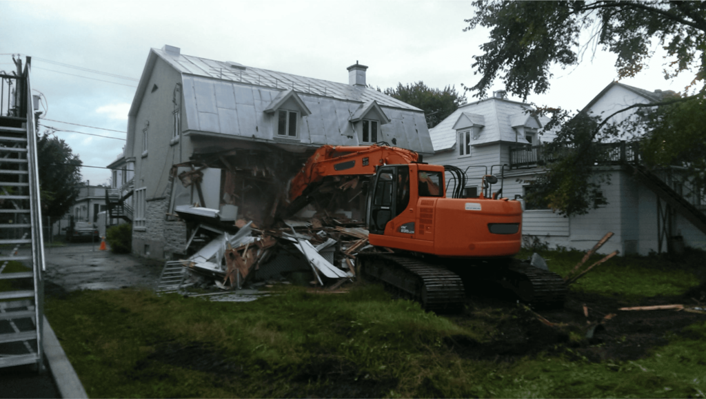 Une excavatrice orange en train de démolir un bâtiment résidentiel à toit métallique, avec des débris de bois et de métal éparpillés au sol.