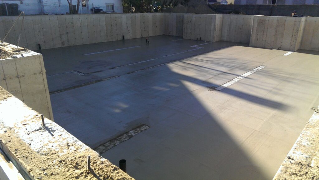 Vue d’une dalle de béton fraîchement coulée à l’intérieur d’un sous-sol en construction, avec murs de fondation en béton et tuyaux de plomberie apparents.