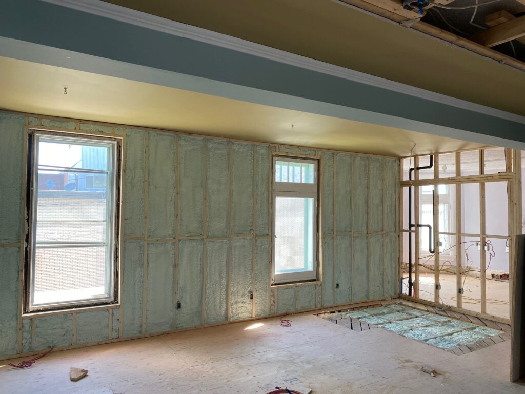 Mur en cours d’isolation avec mousse projetée, fenêtres installées et structure en bois apparente lors d’une rénovation commerciale.
