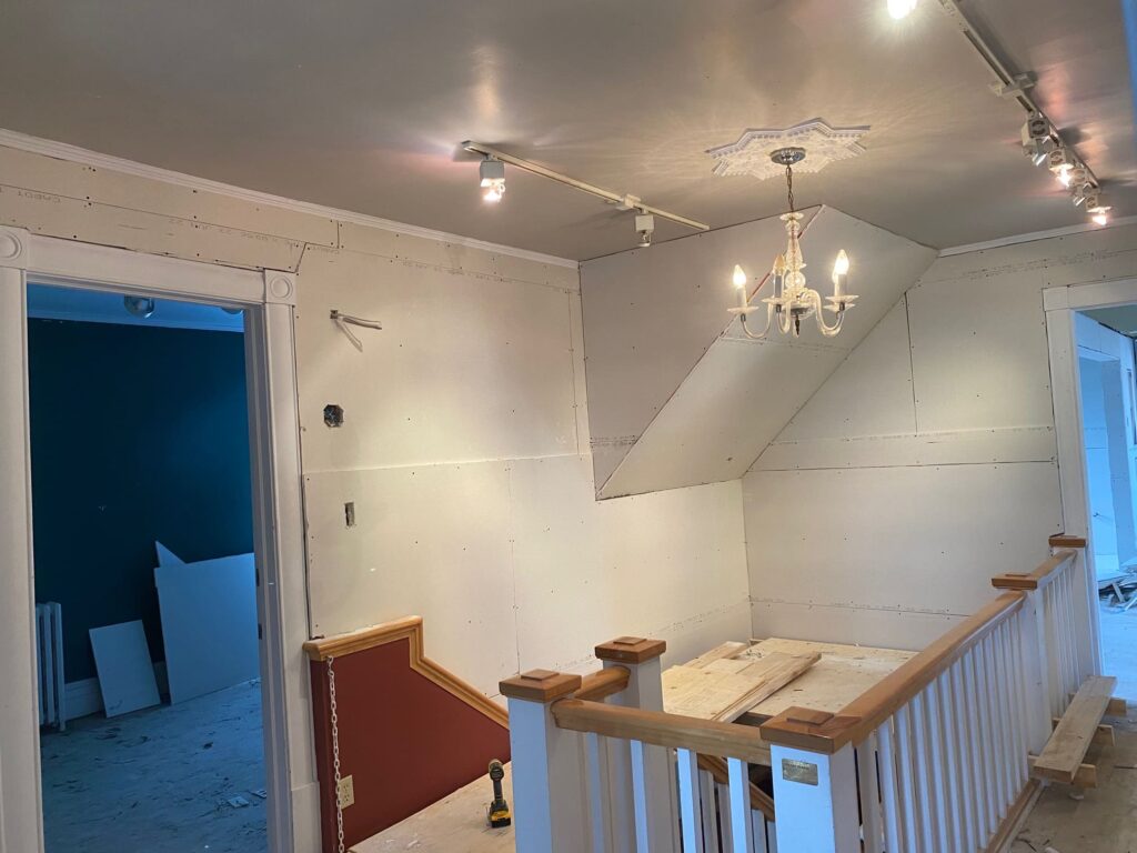 Espace intérieur en rénovation avec des panneaux de gypse installés, un lustre suspendu et une rampe d’escalier en bois.