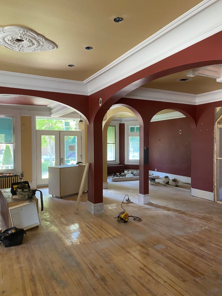 Espace intérieur en rénovation avec arches rouges, moulures blanches et plancher en bois en cours de restauration.